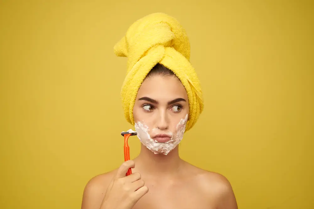 woman with razor i hand and shaving foam on face on yellow background